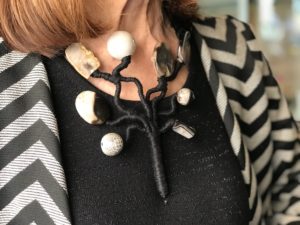 Dettaglio della collana indossata da Cilla. È un pezzo unico realizzato per l’alta moda di grande pregio. Realizzata interamente a mano questa collana riproduce un ramo stilizzato, tutto ricoperto a mano con filato in seta. Ogni ramo termina con un elemento diverso, preziosissimo: luminose ceramiche e preziose pietre dure, splendide agate striate nei toni raffinatissimi del tortora e nero. Un incanto su un abito da sera, di carattere indossata con un tailleur. Un capolavoro di lavorazione artigianale.
