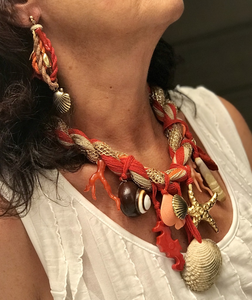 Splendida collana indossata da Sandra, una creazione dalla fine lavorazione artigianale nelle calde nuance del corallo e naturale illuminate dall'oro. Base a cordone, fatto a mano, con filati di cotone e rete in lurex dorata, splendidi ciondoli con elementi a tema marino: conchiglie naturali ed elementi in plexiglas. Orecchini in parure, realizzati con la stessa tecnica e gli stessi materiali. Sono pezzi unici di Cillabijoux.